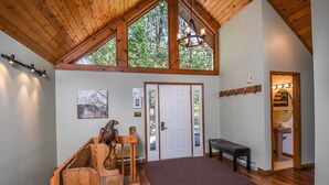 Une pièce spacieuse avec un plafond haut, des poutres en bois, une grande fenêtre, un banc et une porte donnant sur une salle de bain.