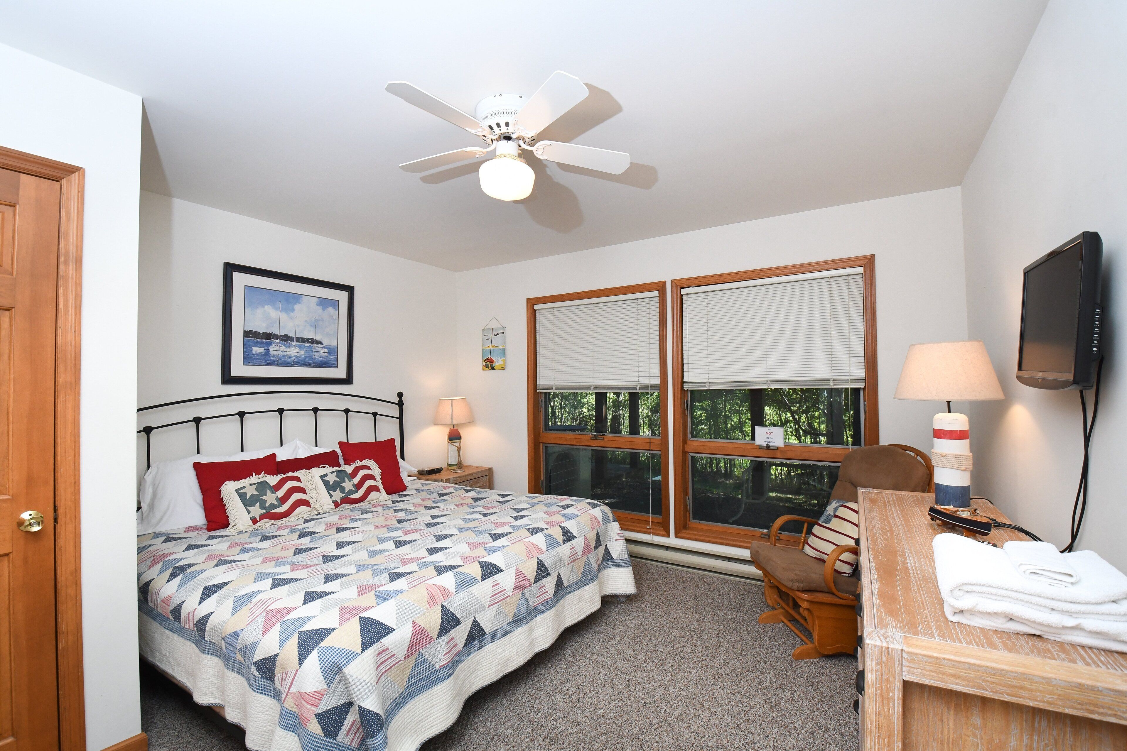 A bedroom with a bed, a desk, a chair, a ceiling fan, a TV, and a window with blinds.