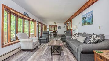 A spacious living room with a grey sofa, striped armchair, and two armchairs. A wooden coffee table is in the center, and there are large windows on one side.