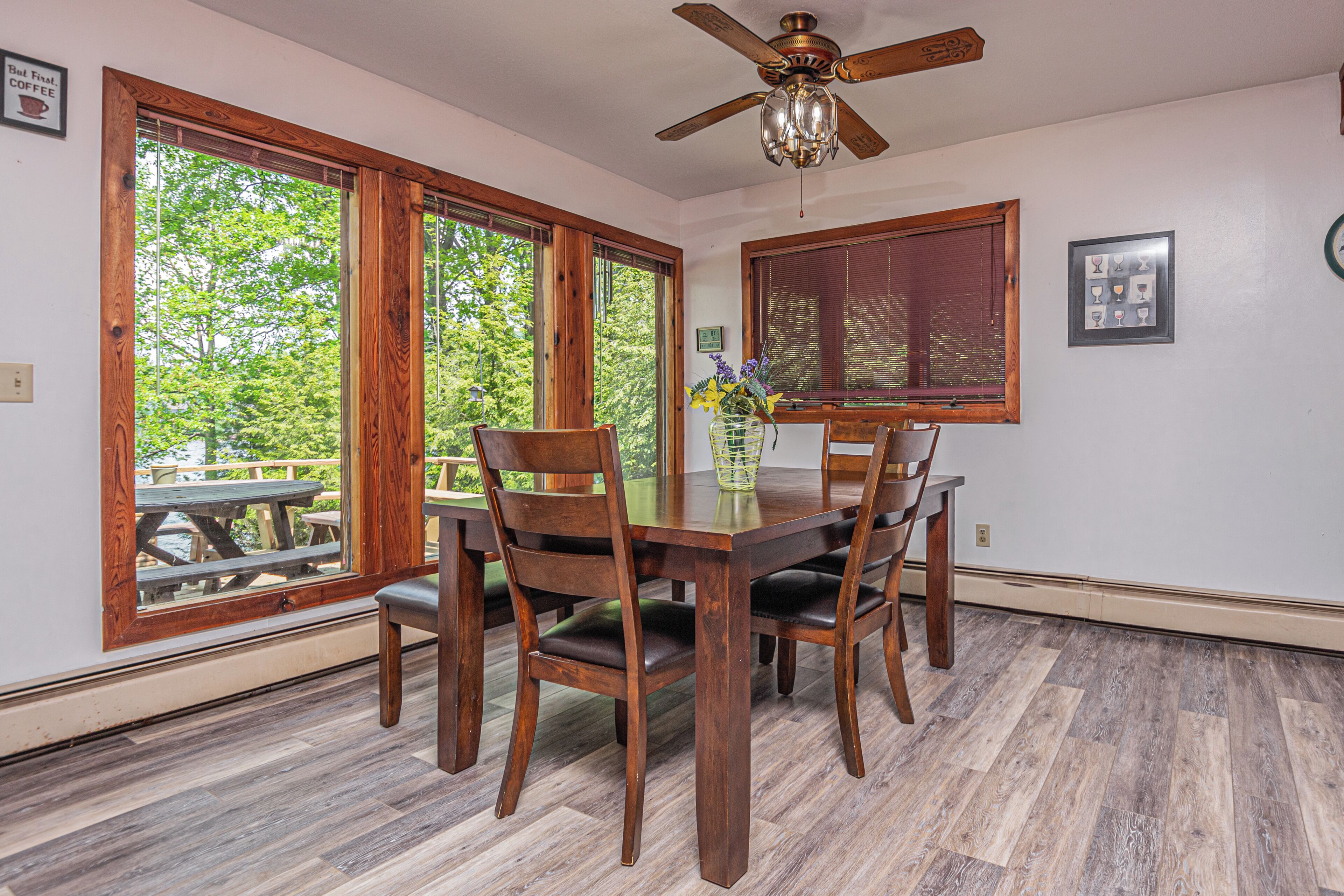 A dining area with a wooden table and chairs, large windows with wooden frames, and a ceiling fan.