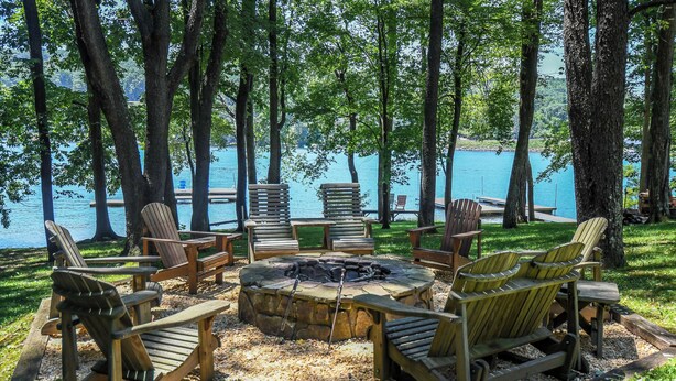 A circular fire pit surrounded by wooden Adirondack chairs, set on a gravel area with a lake view.