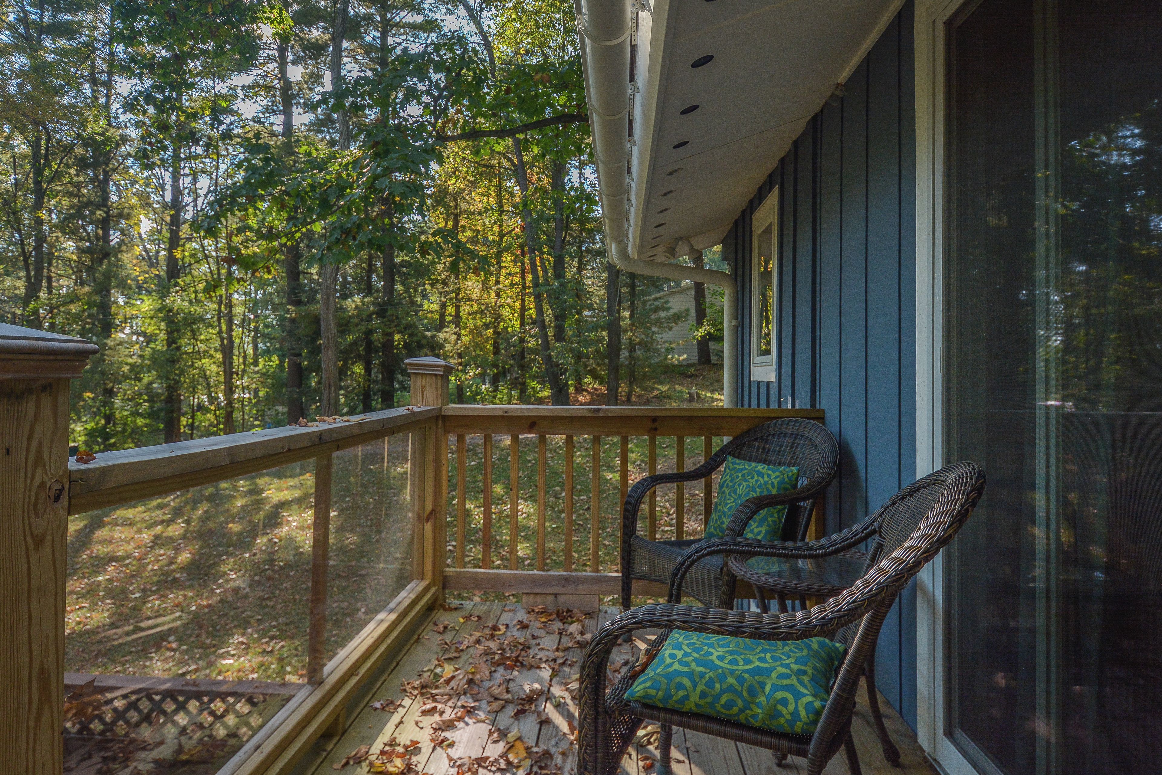 A wooden deck with a railing, two wicker chairs with patterned cushions, and a view of a forested area.