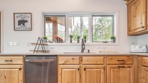A kitchen with wooden cabinets, a stainless steel dishwasher, a window with plants, and a wall sign.