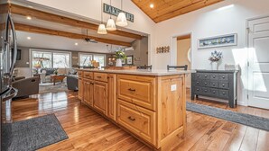 A spacious kitchen with wooden cabinets, a central island, pendant lights, and a view of the living area.