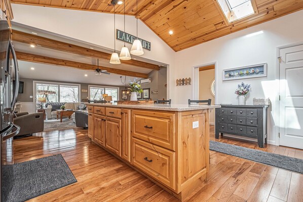 A spacious kitchen with wooden cabinets, a central island, pendant lights, and a view of the living area. - Lakeside Dreamin' by Taylor Made Deep Creek (McHenry)
