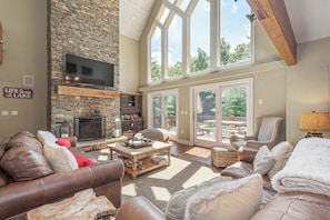 A spacious living room with a stone fireplace, leather furniture, and large windows offering views of the outdoors. - Lakeside Story by Taylor Made Deep Creek (Swanton)