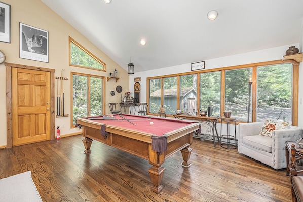 A pool table in a room with large windows, wooden floors, and a cozy seating area.