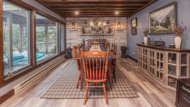 A dining area with a wooden table and chairs, a fireplace, and large windows overlooking a garden. - Lakeside Vista by Taylor Made Deep Creek (Oakland)