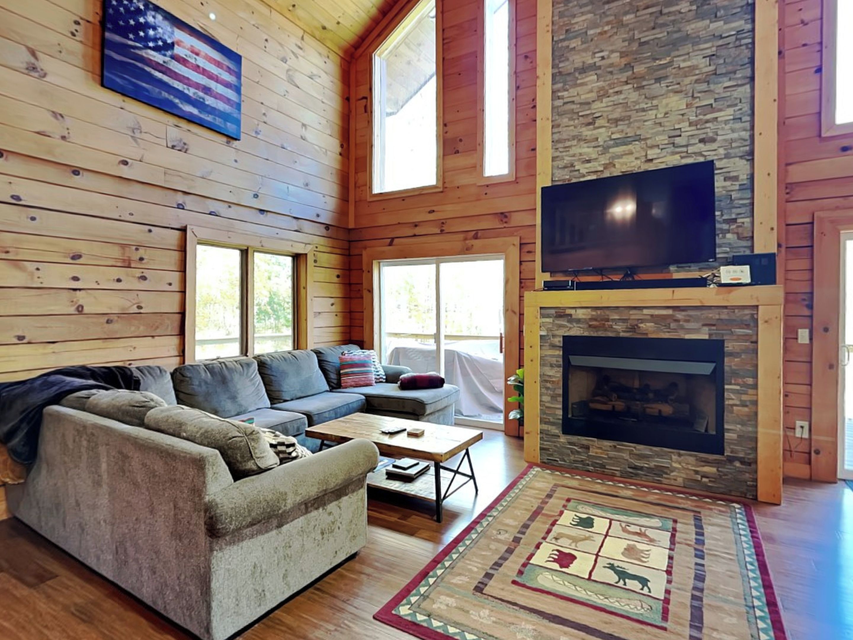 Un salón amplio con un sofá modular, un televisor de pantalla plana sobre la chimenea de piedra y grandes ventanales que dejan entrar luz natural.