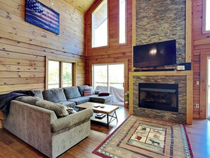 A spacious living room with a sectional sofa, a flat-screen TV mounted above a stone fireplace, and large windows allowing natural light. - Lakestar Chalet by Taylor Made Deep Creek (Oakland)