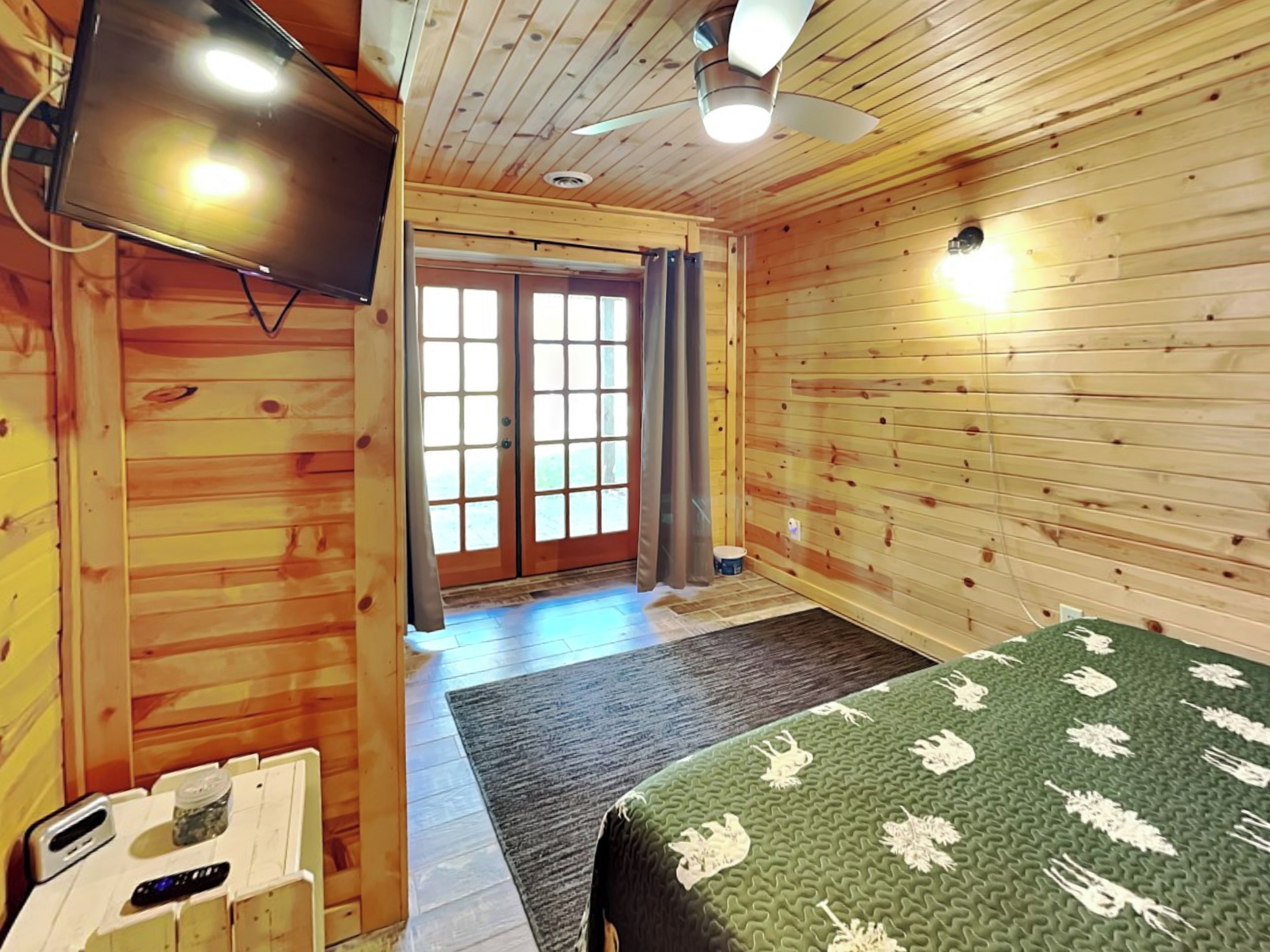 A wooden cabin room with a bed, a television, a ceiling fan, and a sliding glass door.