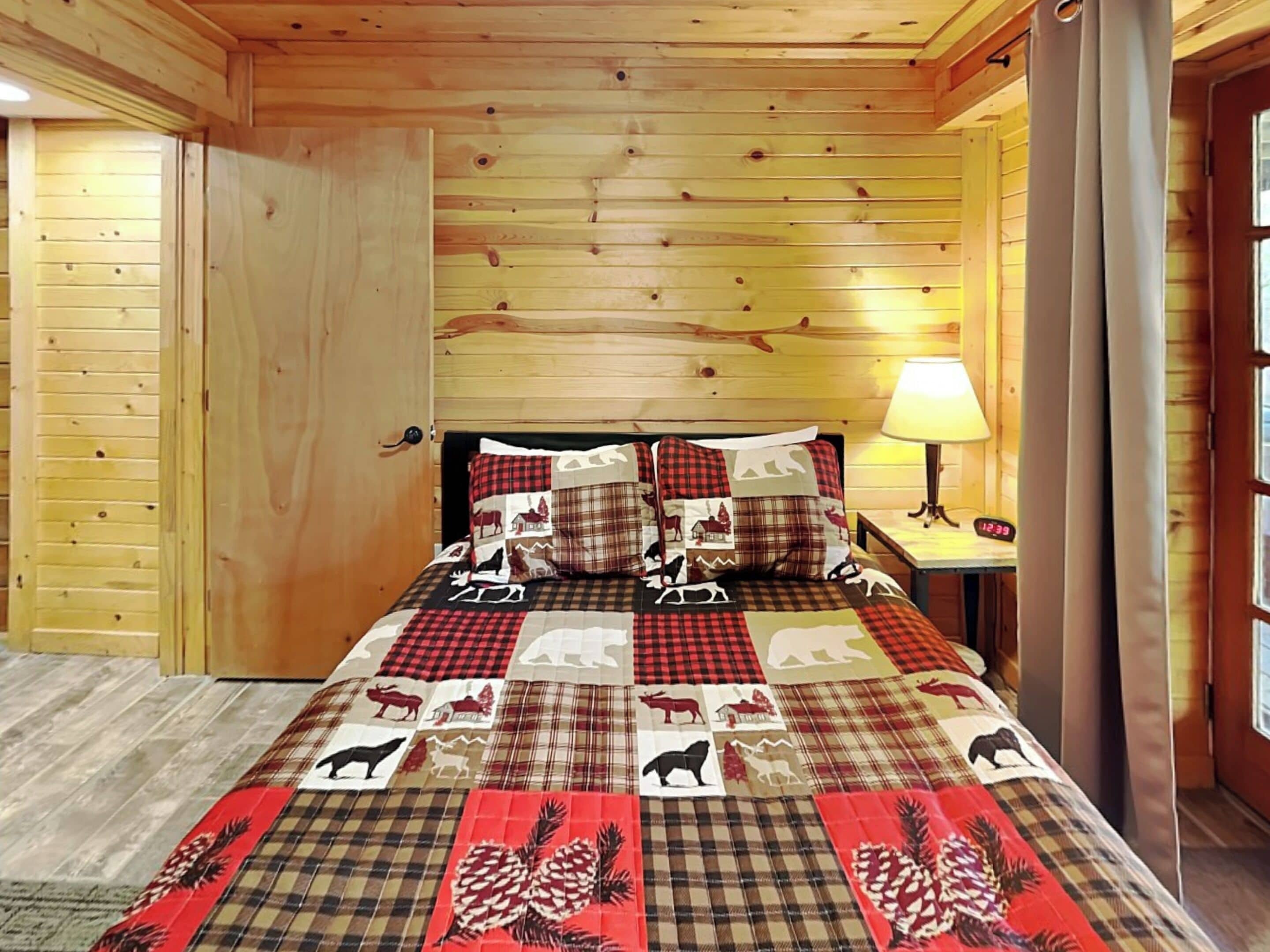 A wooden cabin room with a bed featuring a quilt with animal prints, a nightstand with a lamp, and a wooden door.
