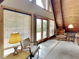 Una sala de estar acogedora con techo de madera, un sofá, un sillón, una mesa con lámpara y ventanas grandes con persianas.