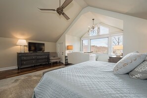 Un dormitorio con una cama grande, ventilador de techo, candelabro, televisor y ventana con vistas.