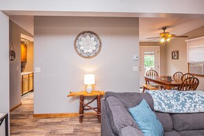 Un salon avec un canapé gris, une table d’appoint en bois sur laquelle se trouve une lampe, un élément décoratif mural et un ventilateur de plafond.