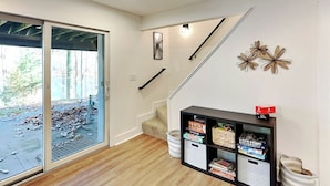 A modern living room with a wooden floor, a sliding glass door leading to an outdoor area, a black entertainment unit with board games, and a staircase with a black railing. - Lazy Days by Taylor Made Deep Creek (McHenry)
