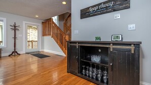 A spacious living area with a wooden floor, a dark wooden cabinet with wine glasses, a staircase, and a sign on the wall. - Leap Away by Taylor Made Deep Creek (Oakland)