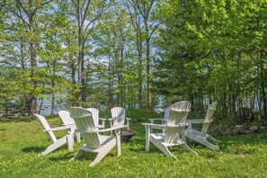 Un grup de cadirees Adirondack disposades al voltant d'una xemeneia exterior a una zona verda, amb arbres i una massa d'aigua al fons.