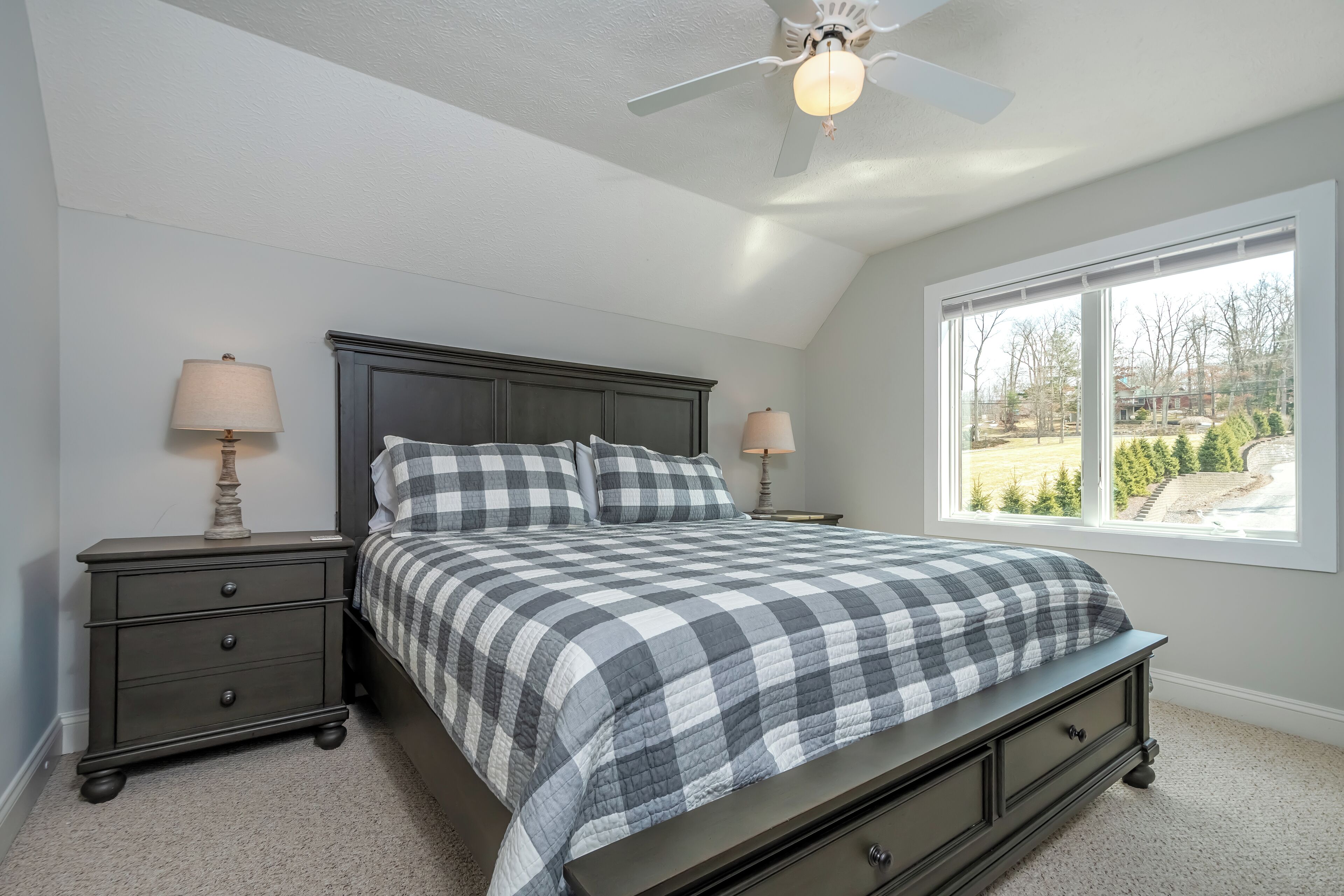 A bedroom with a bed, two nightstands, a ceiling fan, and a window with a view of the outdoors.
