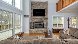 A spacious living room with a stone fireplace, a flat-screen TV, large windows, and a wooden deck visible through the windows. - Leap Away by Taylor Made Deep Creek (Oakland)