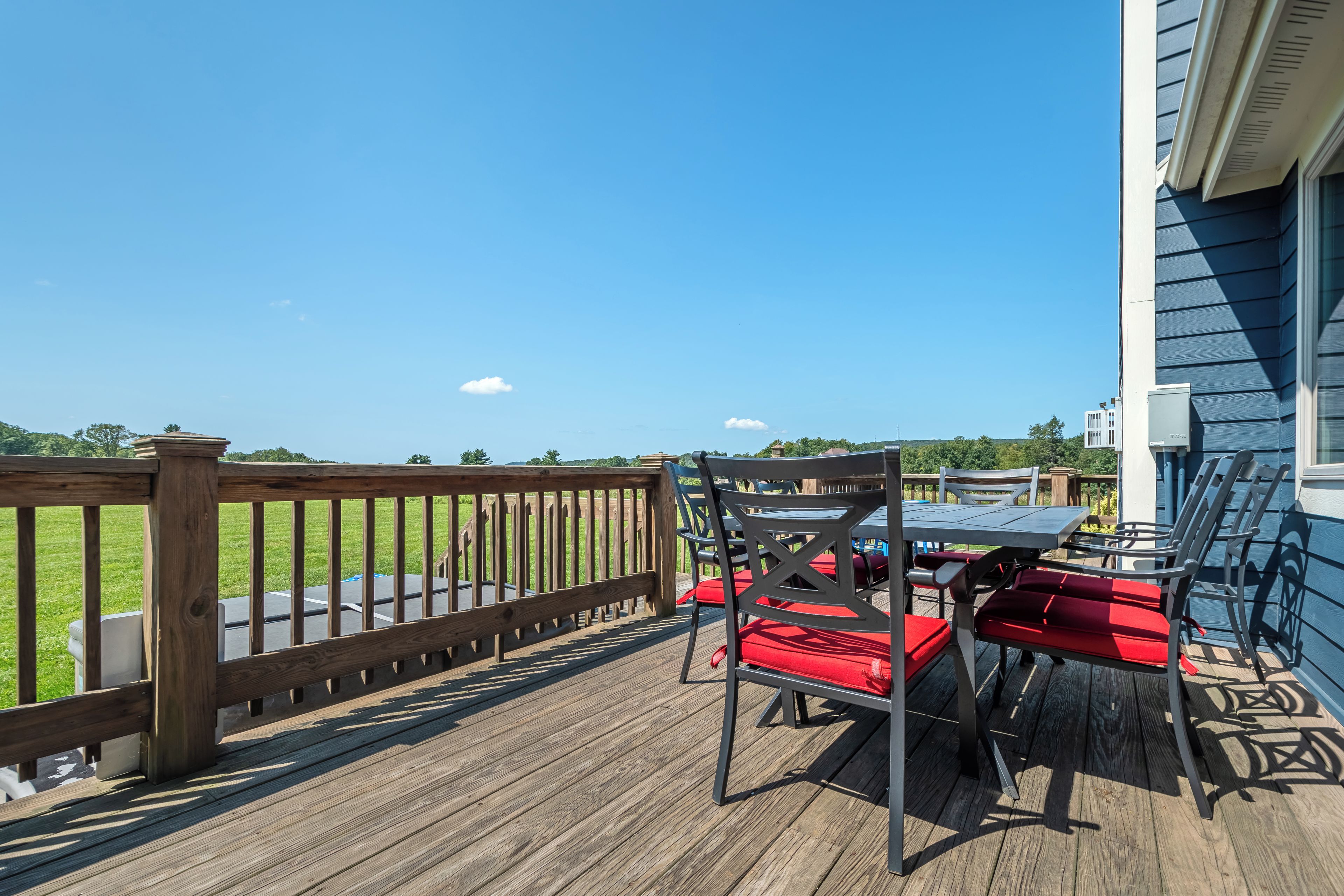 Eine Holzterrasse mit Tisch und Stühlen, Blick auf ein grasbewachsenes Feld, und ein Gebäude mit blauen Verkleidungen.