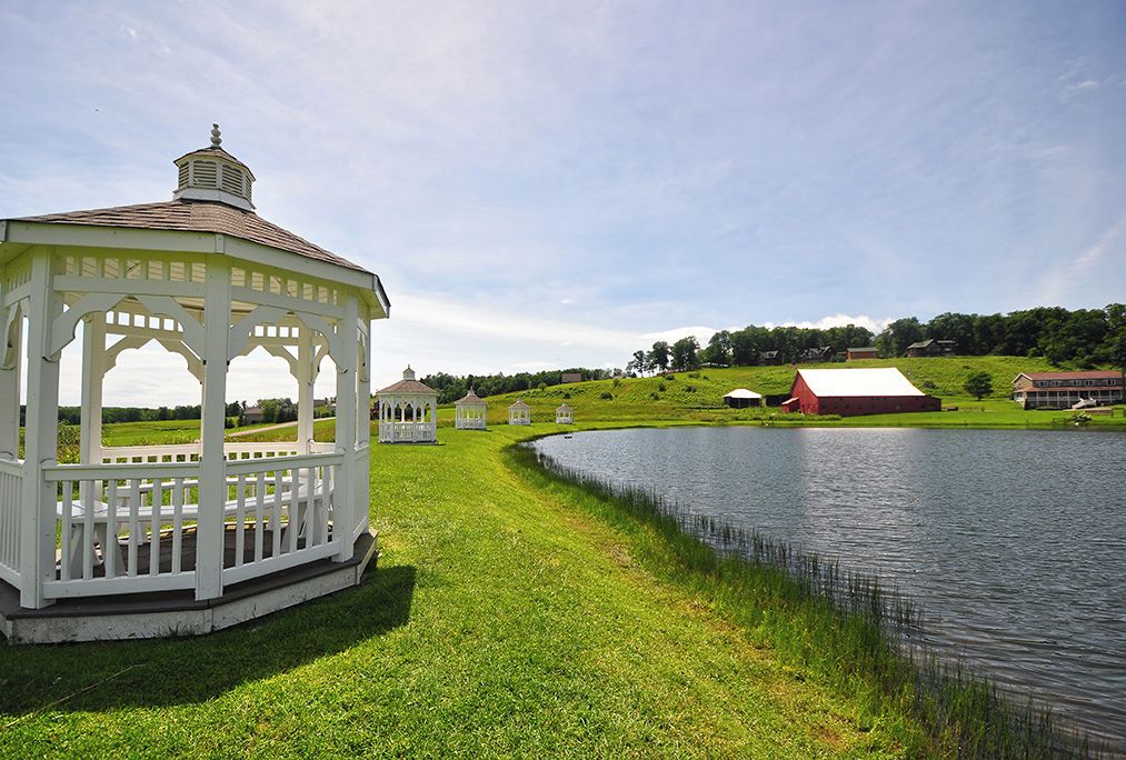 A gazebo with a white roof and open sides, surrounded by a grassy area and a body of water.