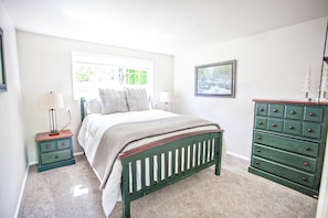 Une chambre à coucher comprenant un lit, deux tables de chevet, une commode et une fenêtre donnant sur un paysage verdoyant.