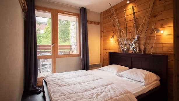 Une chambre à coucher avec une tête de lit en bois, un lit recouvert de linge de lit blanc, une grande fenêtre avec des rideaux et une vue sur l’extérieur.