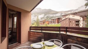 Un balcon avec une table dressée pour deux personnes, donnant sur une région montagneuse où l’on aperçoit des bâtiments au loin.