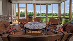 Un patio exterior con una mesa redonda rodeada por seis sillas marrones con cojines rojos, ventanas amplias que ofrecen vistas a un paisaje verde y una chimenea de piedra.