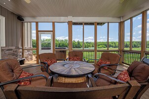 An outdoor patio with a round table surrounded by six brown chairs with red cushions, large windows offering a view of a green landscape, and a stone fireplace.