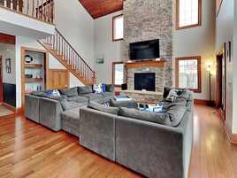 A spacious living room with a large sectional sofa, a stone fireplace, and a wooden staircase.
