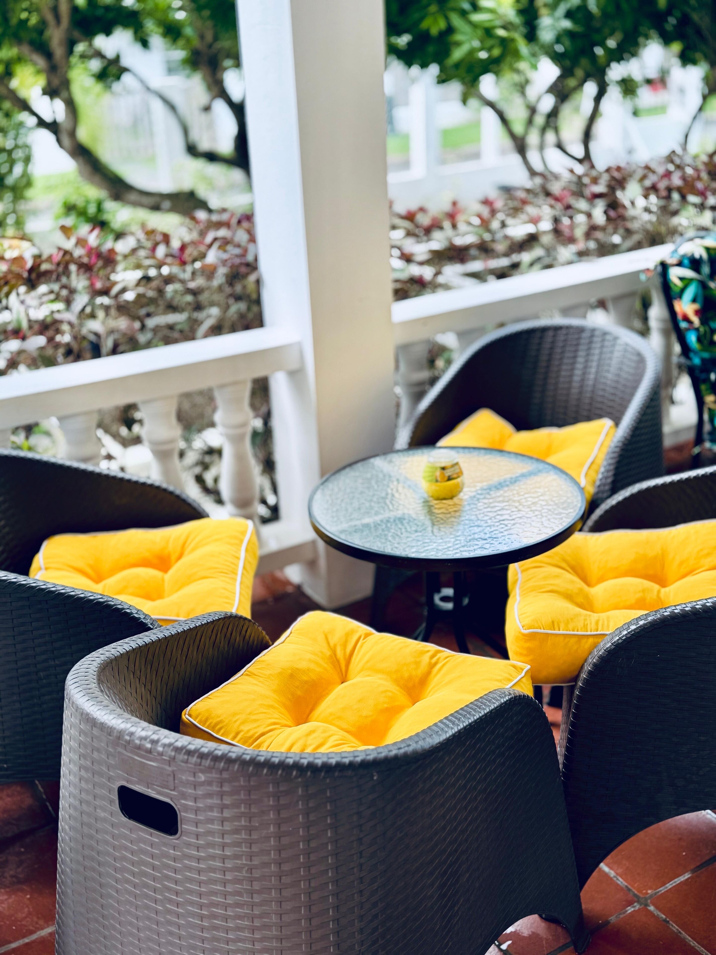 A patio area with wicker chairs and yellow cushions, a small round table with a bottle, and a white railing with greenery in the background.