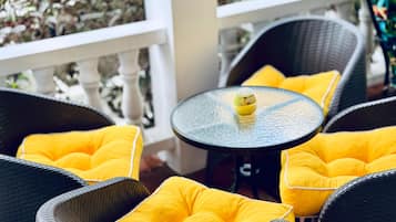 A patio area with wicker chairs and yellow cushions, a small round table with a bottle, and a white railing with greenery in the background.