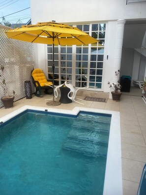 Une piscine dont le bord est recouvert de carreaux blancs et bleus, avec un parasol jaune et une chaise jaune.