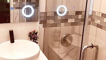 A modern bathroom with a glass-enclosed shower, a white sink with a gold faucet, and a mirror with a circular light.