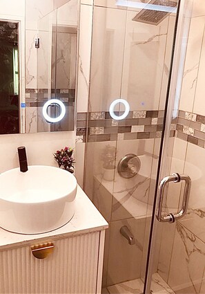 Et moderne badeværelse med en brusekabine i glas, en hvid vask med guldarmatur og et spejl med cirkulær lysring.