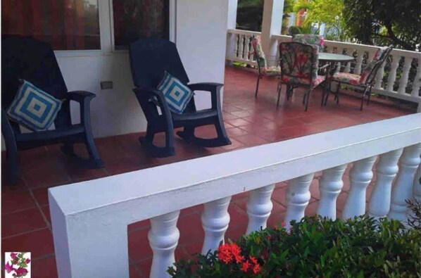 Un espace extérieur abrité avec deux chaises et une table, une balustrade blanche et un pot de fleurs.