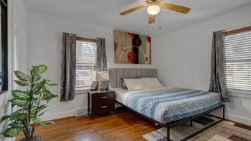 A bedroom with a bed, a nightstand, a ceiling fan, a painting, and a potted plant.
