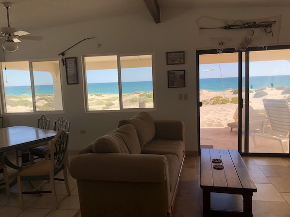 A living room with a beige sofa, a wooden table, and a dining area with chairs. Large windows offer a view of the beach and ocean.