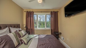 A bedroom with a large bed, a ceiling fan, a window with curtains, and a flat-screen TV mounted on the wall. - Live a Little by Taylor Made Deep Creek (McHenry)