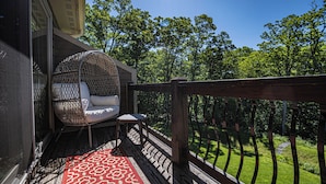 Un espace extérieur accueillant, aménagé avec un fauteuil confortable et une petite table, un tapis rouge à motifs et une balustrade en bois donnant sur un paysage verdoyant.