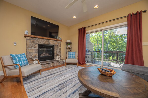 A living room with a stone fireplace, a round wooden coffee table, and a view of the outdoors through large sliding glass doors. - Live a Little by Taylor Made Deep Creek (McHenry)
