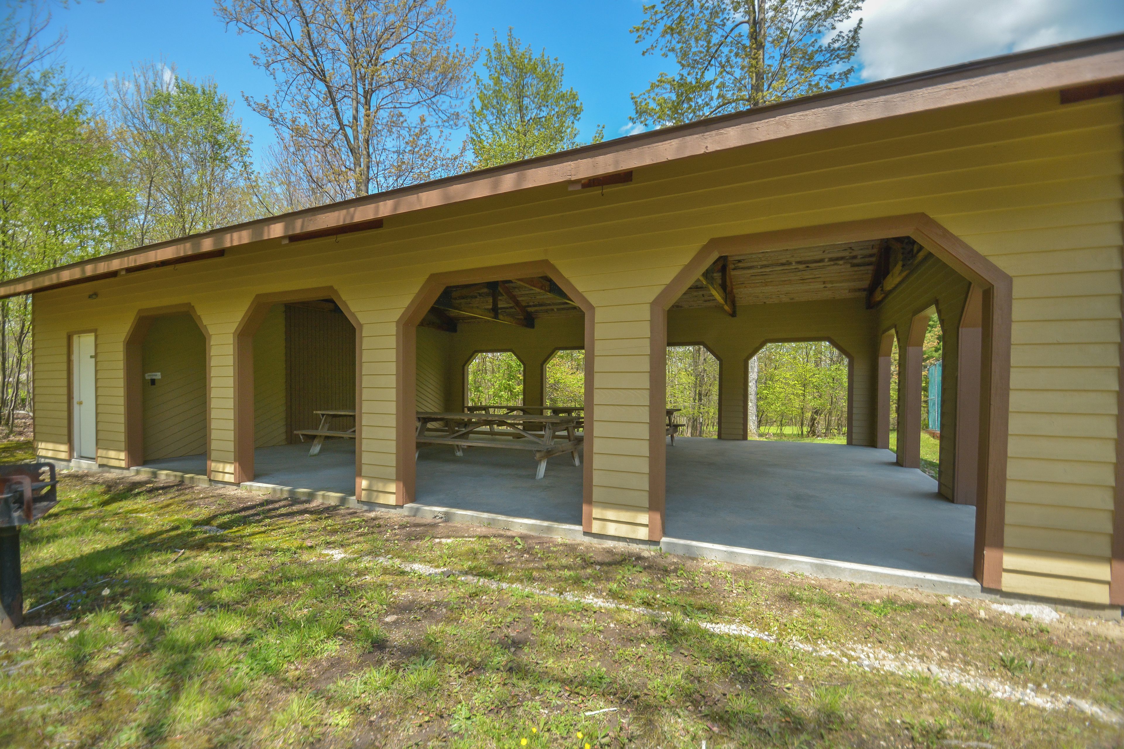 A covered picnic area with benches and a picnic table, surrounded by trees.