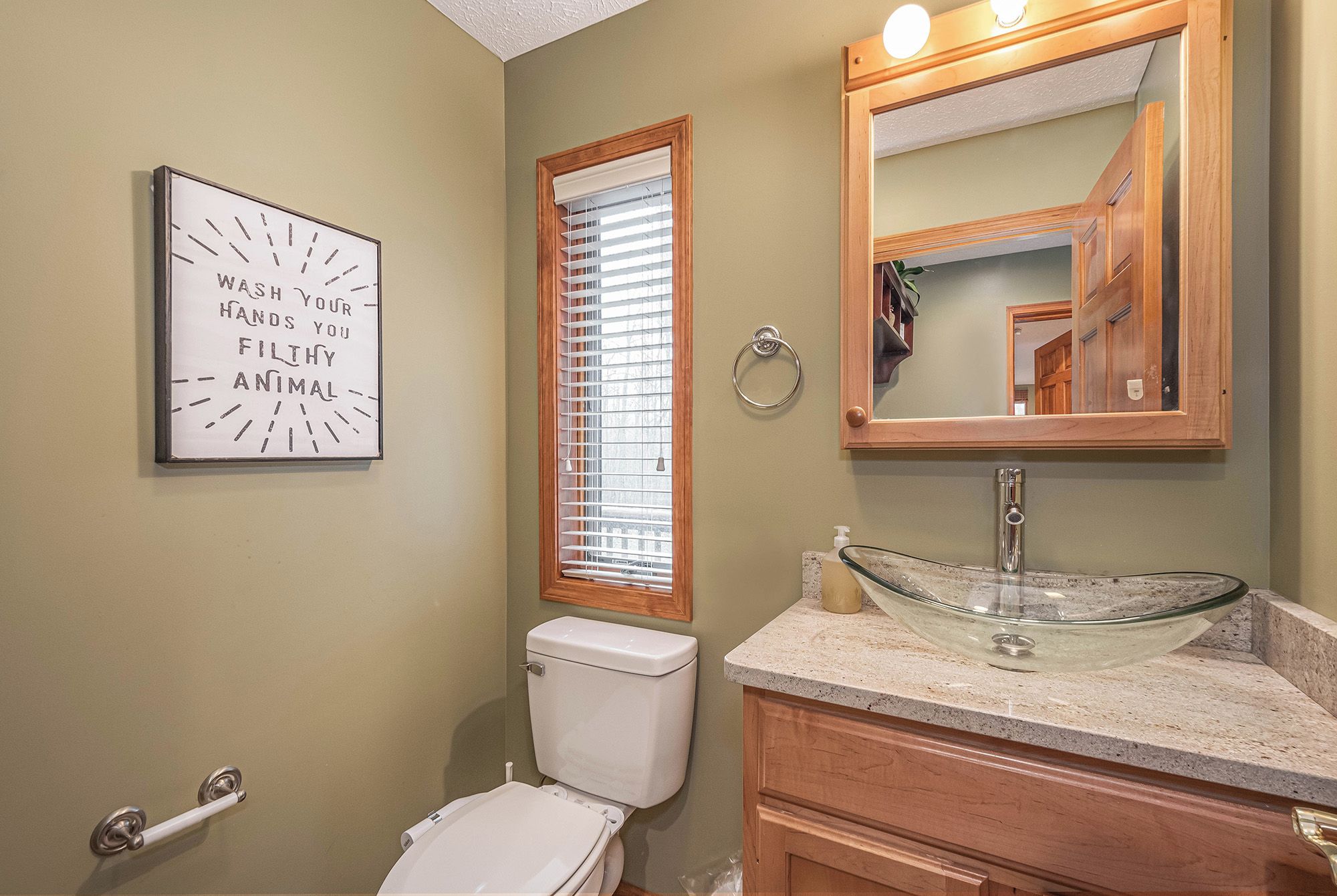 A bathroom with a toilet, a glass vessel sink, a wooden vanity, a mirror, and a window with blinds.