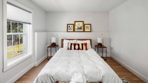 A bedroom with a large window, a bed with white bedding, two bedside tables with lamps, and three framed pictures on the wall.