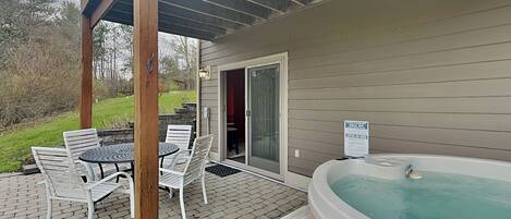 An outdoor patio with a hot tub, a table with chairs, and a covered area with a wooden ceiling.