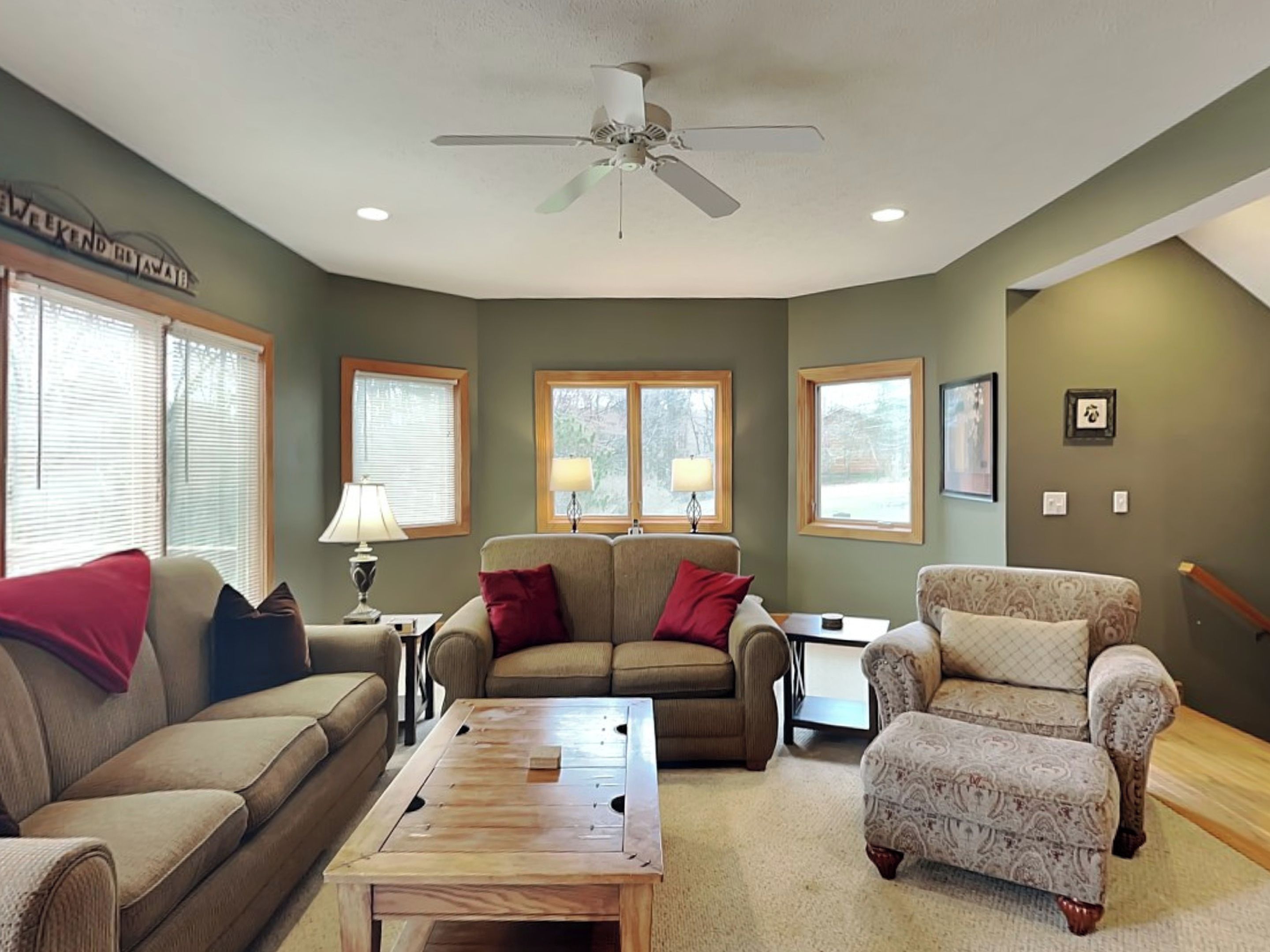 A living room with a ceiling fan, two windows with blinds, a sofa, a loveseat, a patterned armchair, and a coffee table.