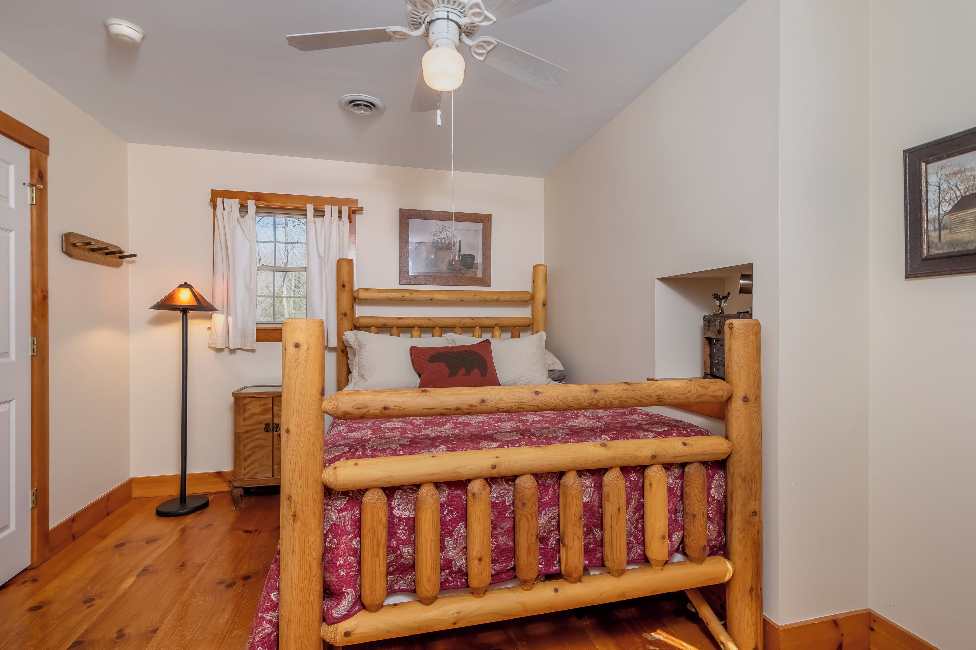A wooden bed with a headboard, a bedside table, a lamp, a window with curtains, and a ceiling fan.