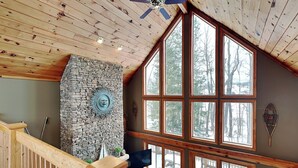 A spacious room with a high ceiling, large windows, a stone fireplace, and a wooden staircase. - Midnight Dip by Taylor Made Deep Creek (Swanton)